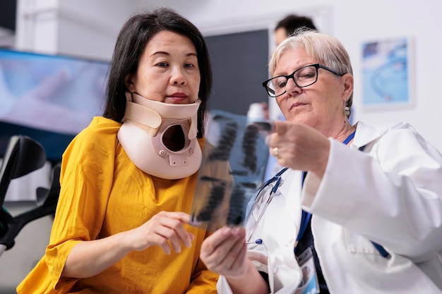 Médico sênior consultando paciente ferido com colar cervical na área de espera do hospital, analisando o exame de radiografia dos pulmões, médico antigo e paciente discutindo sobre saúde na visita de check-up