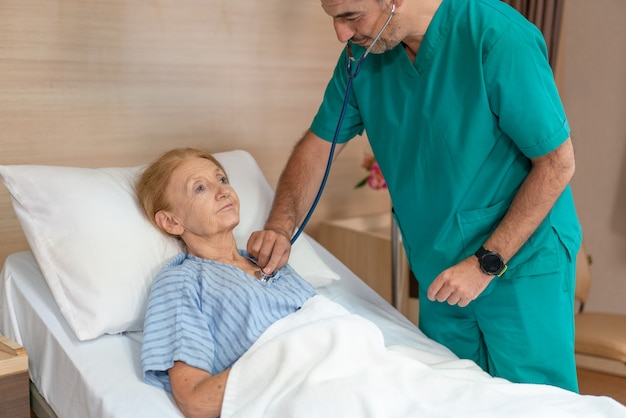 Médico sênior homem ouvindo respiração com estetoscópio para paciente idosa Mulher idosa deitada na cama sorrindo e feliz enquanto é tratada no hospital.