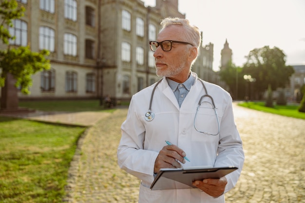 Médico sênior sério de jaleco e óculos segurando uma prancheta, olhando para o lado em pé