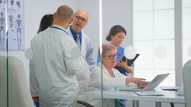 Médico sênior usando laptop explicando aos colegas o tratamento de pacientes durante o brainstorming médico no escritório de reunião no hospital. equipe médica discutindo diagnóstico de registro de problemas no local de trabalho.