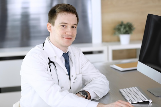 Médico sentado atrás de um computador no local de trabalho na clínica ou no hospital Fotografia na cabeça do médico e conceito de medicina