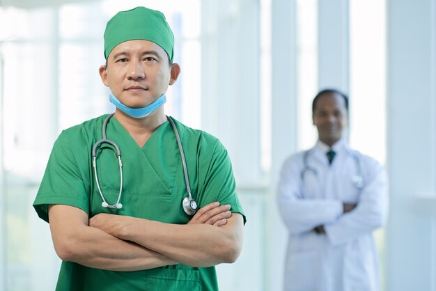 Médico sério e confiante em uniforme verde cruzando os braços e olhando para a câmera