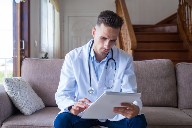 Médico de sexo masculino joven enfocado en el sofá y bata médica trabajando con el libro de registro en papel, tomando notas, administrando las visitas de los pacientes, registrando el tratamiento médico, sentado en el sofá en casa.