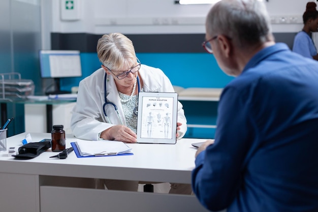 Médico sosteniendo una tableta con la imagen del esqueleto humano para explicar el diagnóstico al anciano en el gabinete. Médico y paciente que miran la ilustración de los huesos y la médula espinal en el dispositivo para su examen.