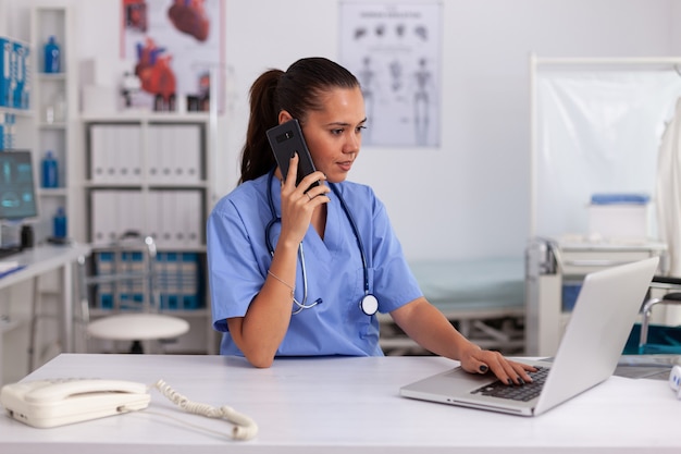 Médico con teléfono y portátil en la oficina del hospital vistiendo uniforme azul