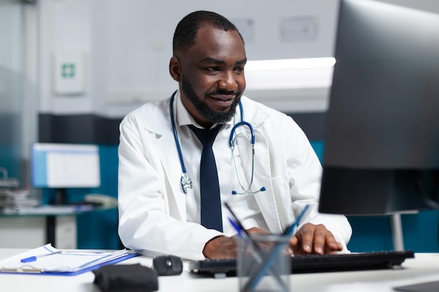 Médico terapeuta afro-americano de uniforme, analisando a experiência da doença na prescrição de remédios on-line de digitação de computador. Médico homem monitorando relato do paciente de sintomas no consultório do hospital