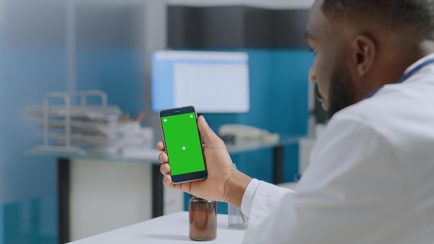 Médico terapeuta afroamericano sosteniendo simulacros de teléfono con pantalla verde chroma key con pantalla aislada analizando informe de enfermedad. Hombre médico que trabaja en el tratamiento sanitario en la oficina del hospital