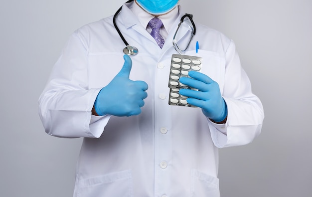 El médico terapeuta está vestido con una bata blanca uniforme y guantes estériles azules están de pie y sosteniendo una pila de píldoras en blister