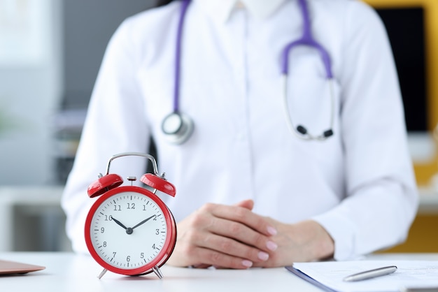 Foto médico terapeuta sentado no escritório em frente a closeup de despertador vermelho. horários médicos