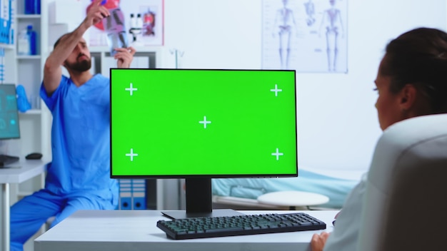Médico trabalhando no computador com tela verde no hospital e assistente olhando para o raio-x. Área de trabalho com tela de maquete substituível na clínica médica enquanto o médico verifica a radiografia do paciente para diagnóstico