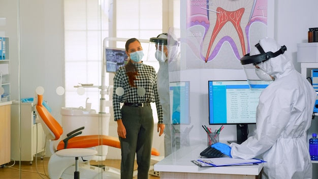 Médico con traje de protección que invita al próximo paciente a la sala de estomatología para el examen de los dientes durante el coronavirus. Médico asistente y dentista vistiendo mono, protector facial, máscara, guantes en clínica dental