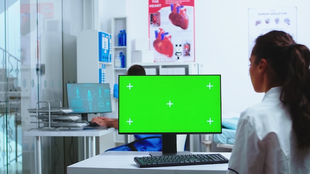Médico usando computador com maquete de tela verde no hospital e assistente sai do armário vestindo uniforme azul. Médico de jaleco branco trabalhando no monitor com chroma key no gabinete da clínica para verificar pa