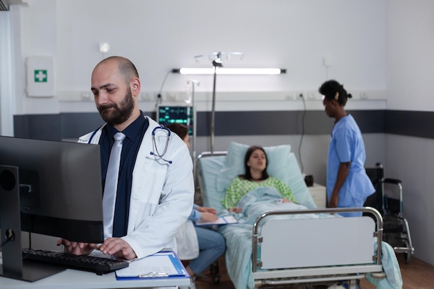 Médico usando computador pessoal para completar o quadro de admissão do paciente enquanto enfermeira e médico conversam com o paciente na enfermaria do hospital. médico com estetoscópio olhando resultados de laboratório na tela.