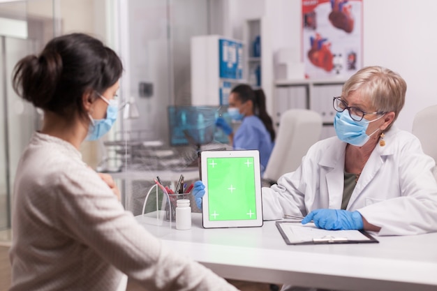 Médico usando máscara de proteção contra coronavírus, olhando para o tablet pc com tela verde durante a consulta ao paciente no consultório do hospital.