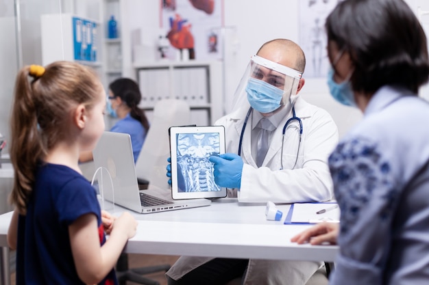 Médico usando máscara facial contra coronavírus explicando a radiografia para mãe e filho durante a consulta