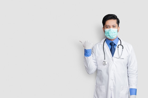 Médico usando máscara uniforme sorrindo enquanto apresenta e aponta isolado na parede branca com espaço de cópia