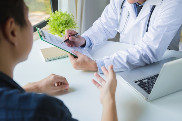 Médico usando un portapapeles para completar un historial médico de la medicación de un joven médico y paciente discutiendo los resultados de un examen físico en una clínica