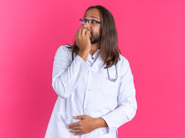 Médico varón adulto ansioso vistiendo bata médica y estetoscopio con gafas manteniendo la mano en el vientre mordiendo los dedos mirando hacia arriba aislado en la pared rosa