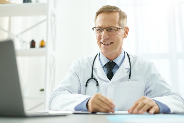 Médico varón alegre con portátil moderno en el trabajo