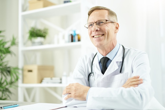 Médico varón alegre sentado en la mesa en el trabajo