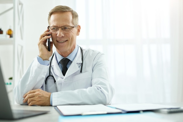 Médico varón alegre tener conversación telefónica en la clínica