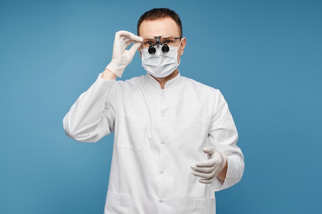 Médico varón caucásico en una mascarilla quirúrgica, guantes protectores y lupas binoculares en el fondo azul, aislado con espacio de copia para texto o colocación de productos