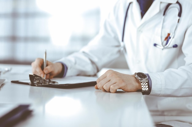 Médico varón desconocido sentado y trabajando con el portapapeles del registro del historial de medicamentos en la clínica en su lugar de trabajo primer plano Médico joven en el trabajo Concepto de medicina de servicio médico perfecto