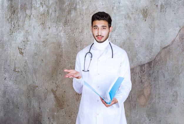 Médico varón con estetoscopio sosteniendo una carpeta azul e interactuando con la persona que lo rodea.