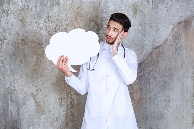 Foto médico varón con estetoscopio sosteniendo un tablero de información de forma de nube en blanco y parece pensativo.