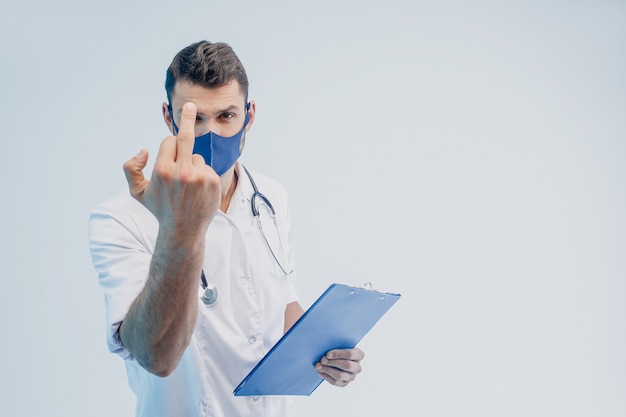 Foto médico varón europeo en máscara protectora muestra el dedo medio. portapapeles de retención joven. persona con estetoscopio con bata blanca. fondo gris con luz turquesa. sesión de estudio. copie el espacio.