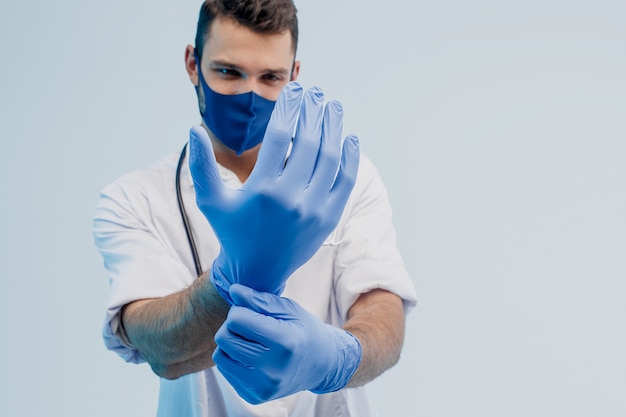 Foto médico varón europeo en máscara protectora poniendo guantes de látex en las manos. hombre joven con estetoscopio con bata blanca. aislado sobre fondo gris con luz turquesa. sesión de estudio. copie el espacio.