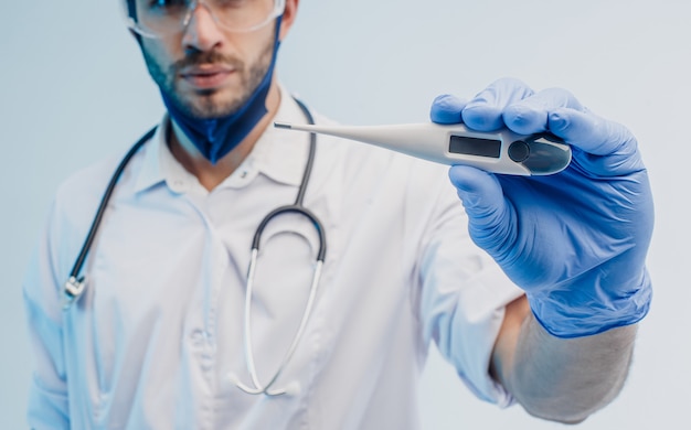 Médico varón europeo que muestra el termómetro digital de cerca. Joven barbudo con estetoscopio con bata blanca, gafas y guantes de látex. Fondo gris con luz turquesa. Sesión de estudio