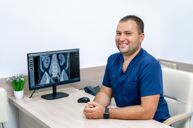 Médico varón guapo joven sentado en la oficina. Consulta médica en consultorio. Fondo de oficina de hospital moderno. Monitor con resultado de resonancia magnética.