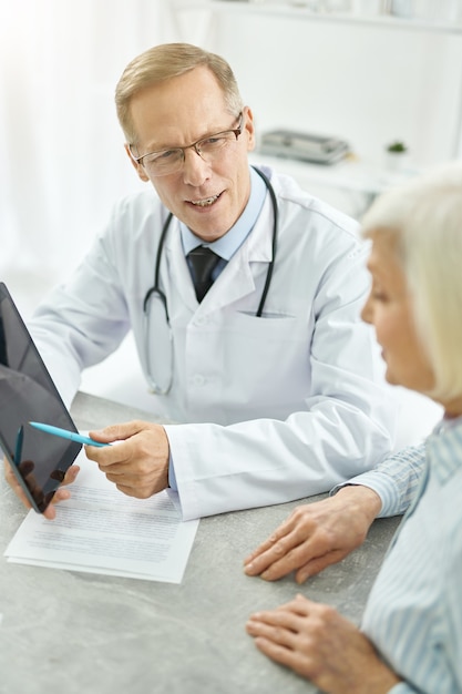 Médico varón guapo sentado en la mesa y apuntando a la almohadilla electrónica PC mientras consulta a una anciana en su oficina