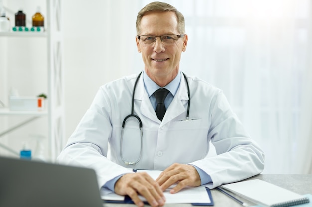 Médico varón guapo sentado en la mesa con documentos en el trabajo