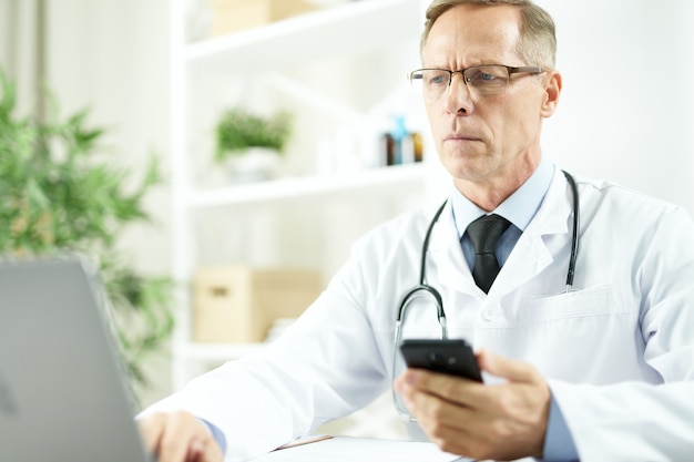 Médico varón guapo con smartphone en sus manos sentado en la mesa y trabajando en el portátil