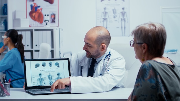 Médico varón joven que explica cómo evitar problemas de huesos a anciana jubilada. Radiología y radiografía en un moderno hospital o clínica privada con personal médico caminando en segundo plano, enfermera