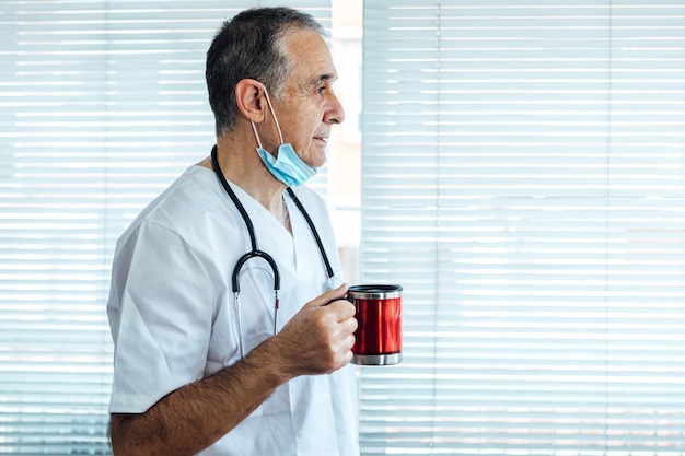 Médico varón maduro - enfermera con máscara en la barbilla tomando café en una ventana del hospital. Covid-19 y concepto de medicina