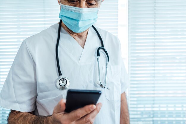 Médico varón maduro - enfermera con mascarilla, mirando el teléfono móvil junto a una ventana del hospital. Covid-19 y concepto de medicina