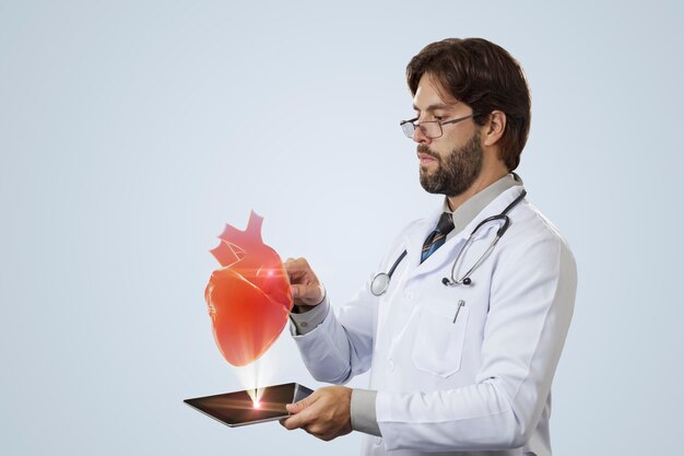 Médico varón mirando un corazón virtual que sale de una tableta en una pared gris