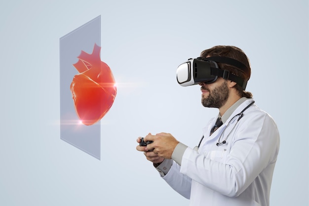 Médico varón en una pared gris con gafas de realidad virtual, mirando un corazón virtual