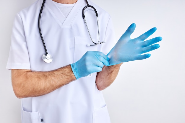 Médico varón recortada lleva guantes preparándose para el trabajo, vestido con bata blanca, aislado en estudio con fondo blanco.