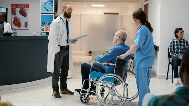 Médico varón reunido con un paciente anciano en silla de ruedas en el vestíbulo de recepción, conversando sobre atención médica y apoyo. Hombre con discapacidad crónica que tiene una visita de control con un especialista.