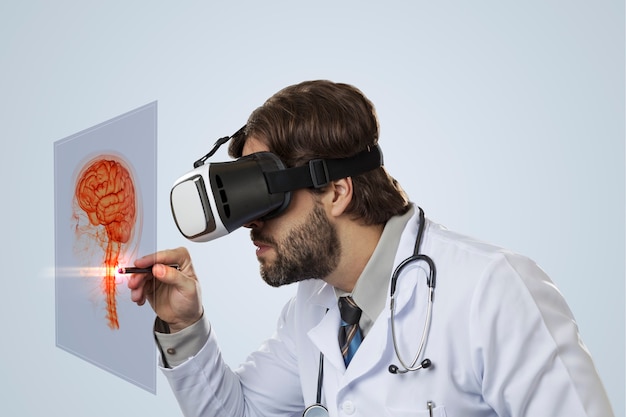 Médico varón sobre una pared gris, utilizando unas gafas de realidad virtual, mirando un cerebro virtual