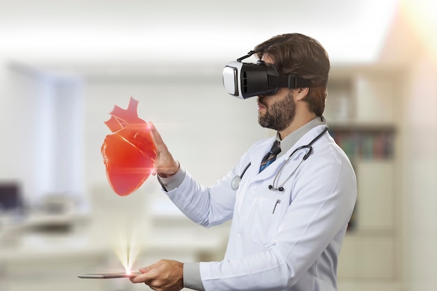 Médico varón en su oficina, utilizando unas gafas de realidad virtual, mirando un corazón virtual