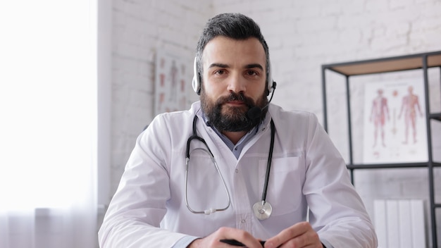 Médico varón vestido con bata blanca que consulta al paciente de forma remota en línea con auriculares. Vista de cámara web.