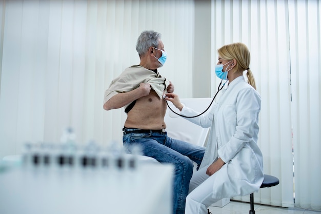 Médico, verificando o sistema respiratório e os pulmões do velho com estetoscópio no escritório do hospital durante a pandemia do vírus corona.