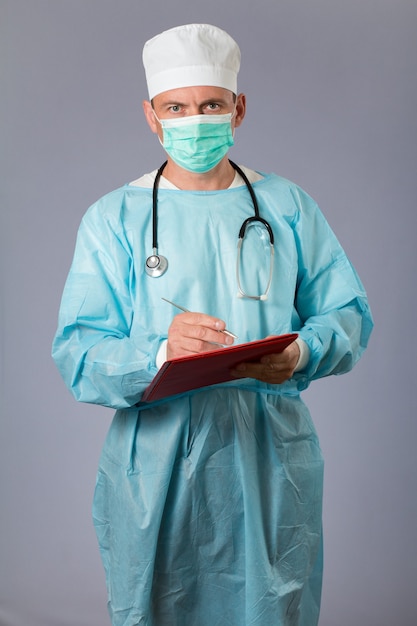 Médico vestido con una bata médica con estetoscopio y mascarilla sosteniendo una pluma y escribiendo en un cuaderno. Fondo gris