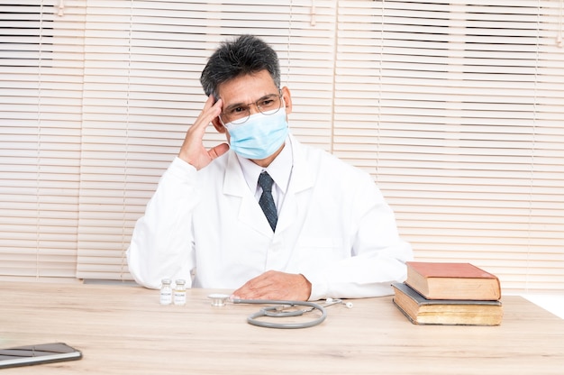 Un médico, vestido con una máscara y ropa adhesiva blanca, se sienta en su escritorio.