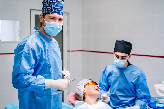 Médico vestindo jaleco azul e máscara convidando você para o consultório dentista e olhando para a câmera com cadeira estomatológica e paciente no.